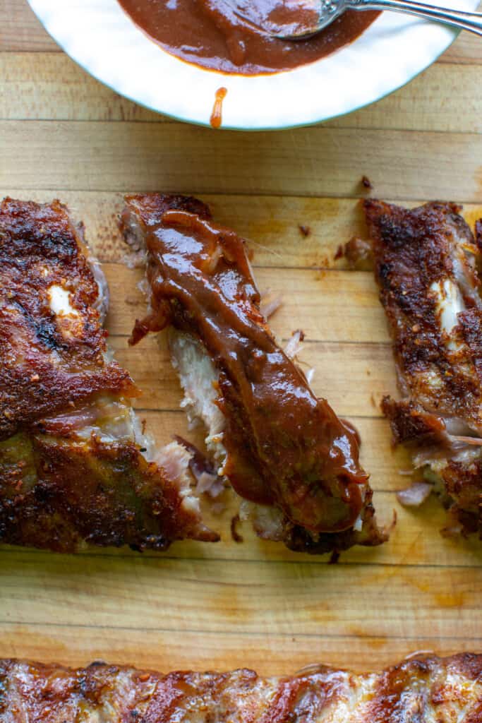 Three ribs sitting on a wood cutting board. One is topped with barbecue sauce sitting next to a white bowl with sauce in it. 