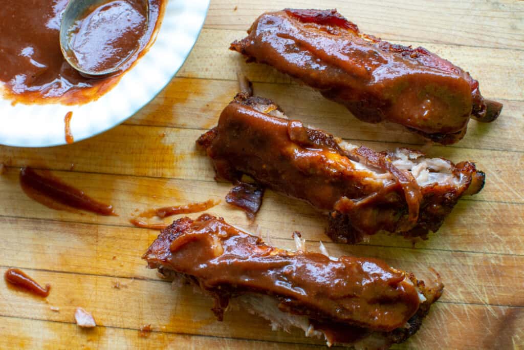 Three individual ribs coated in barbecue sauce sitting on a wood cutting board with a white bowl of sauce. 