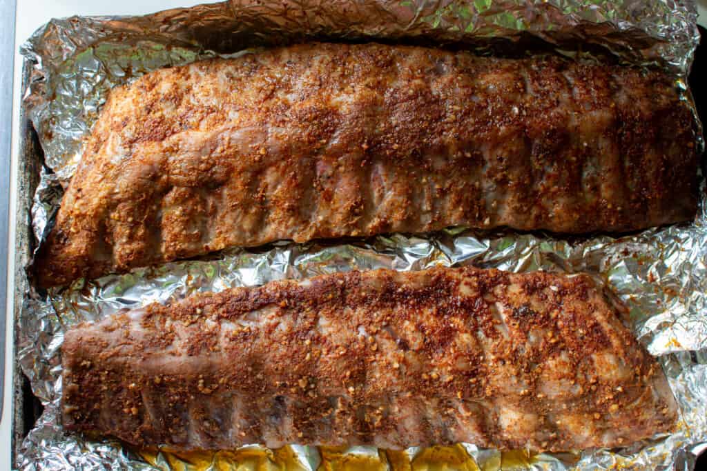 Two racks of ribs sitting on a baking sheet covered with foil that are getting ready to be baked. 