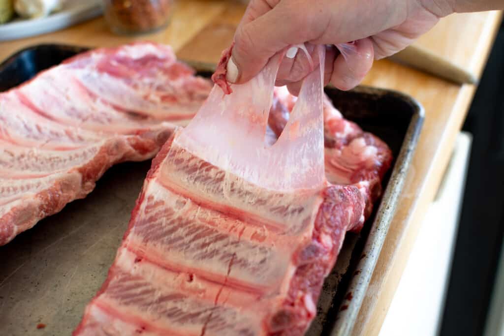 Two racks of baby back ribs sitting on a baking sheet. A hand is pulling a membrane off the back of one of the ribs. 