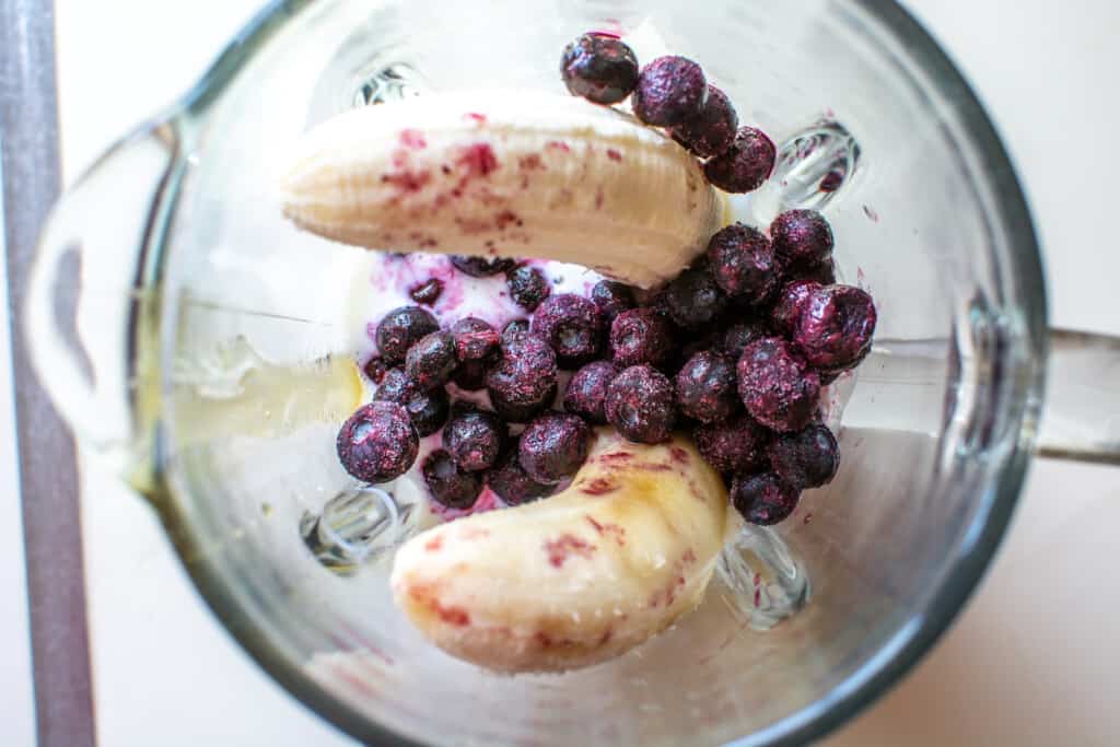 All the ingredients to make banana popsicles in a glass blender including frozen blueberries, bananas, buttermilk, and honey. 