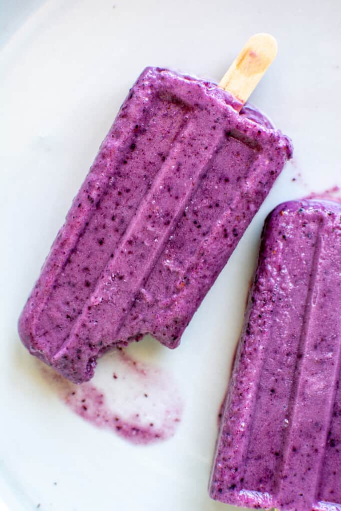 A white plate with two purple popsicles on it and a bite taken out of one of the popsicles. 