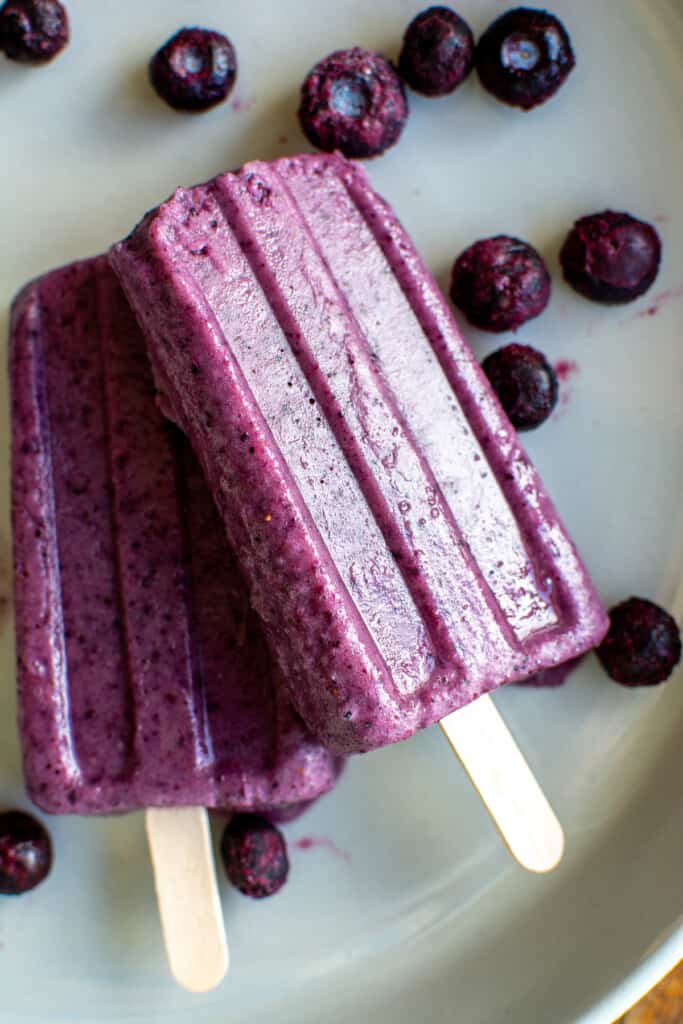 Two banana popsicles on a white plate surrounded by frozen blueberries. 
