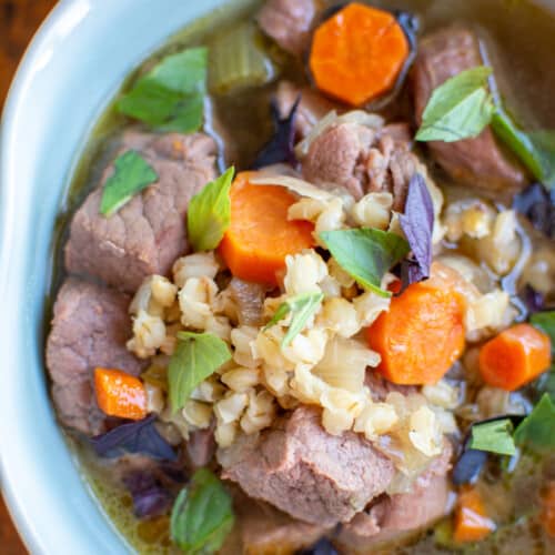 A ceramic bowl of beef and barley soup with hunks of beef, carrots, barley, and fresh herbs.