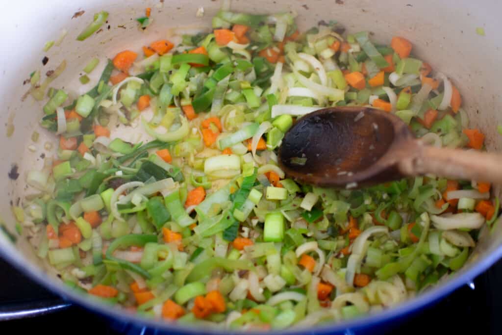 Leeks, carrots, and garlic sautéing in a blue pot with a wood spoon. 