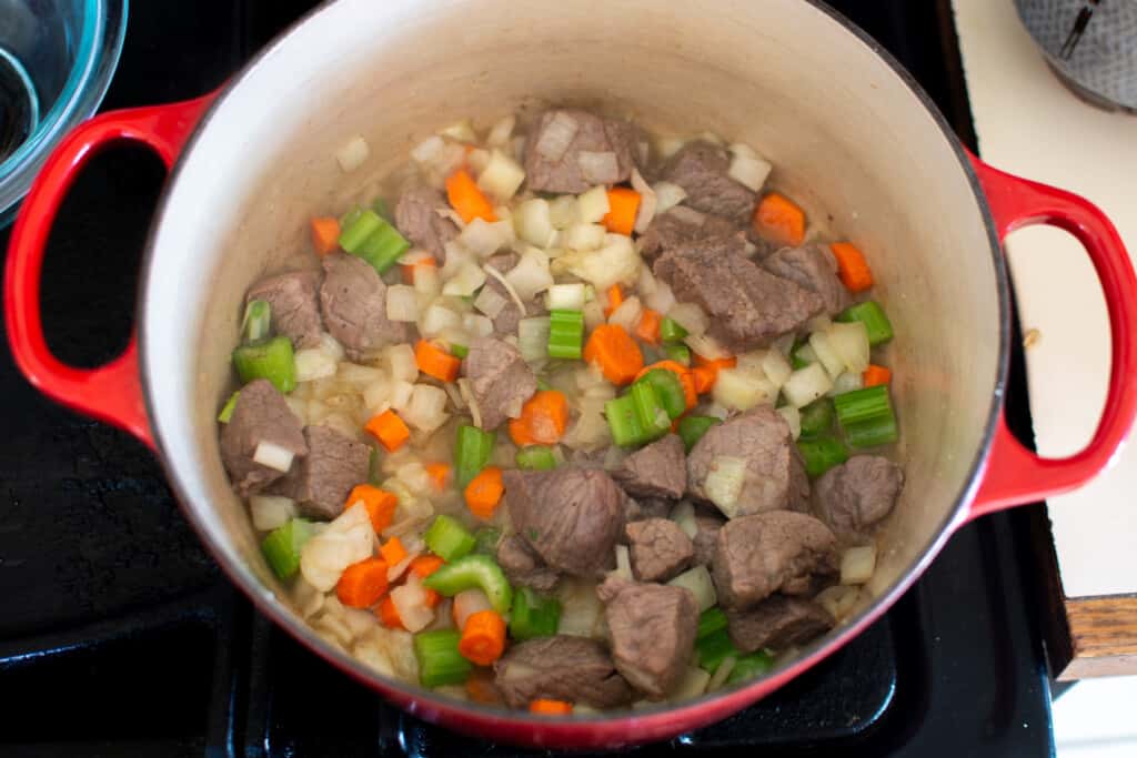 Hunks of browned beef in a pot with red handles plus onions, carrots, and celery in the pot with it. 