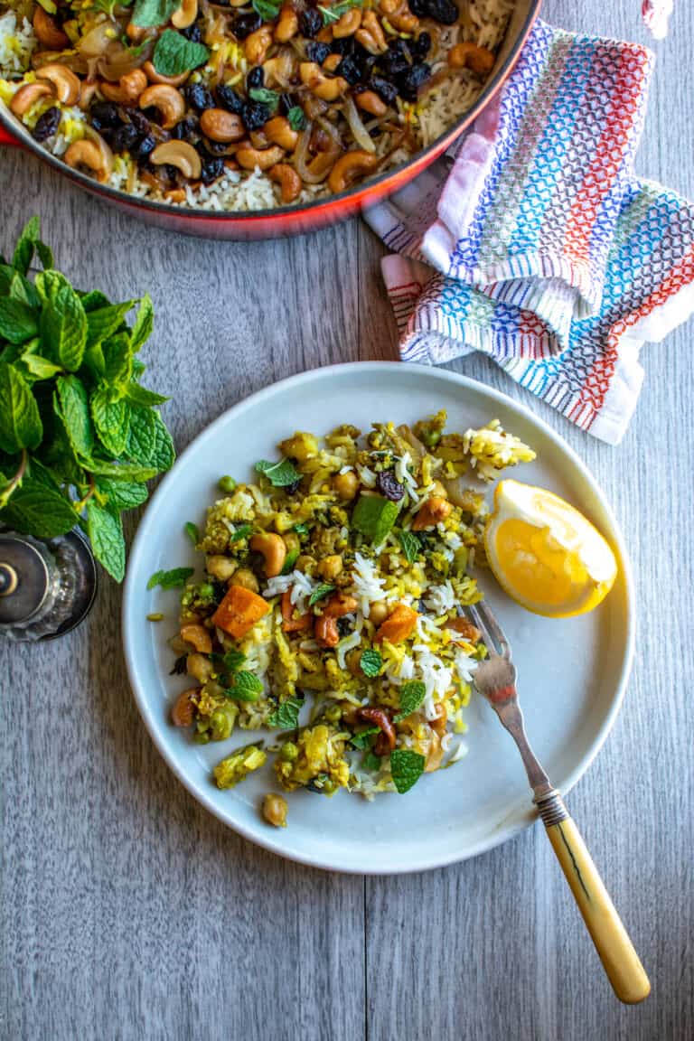 A white plate with vegetable biryani on it sitting on a gray table next to a red pot filled with biryani.
