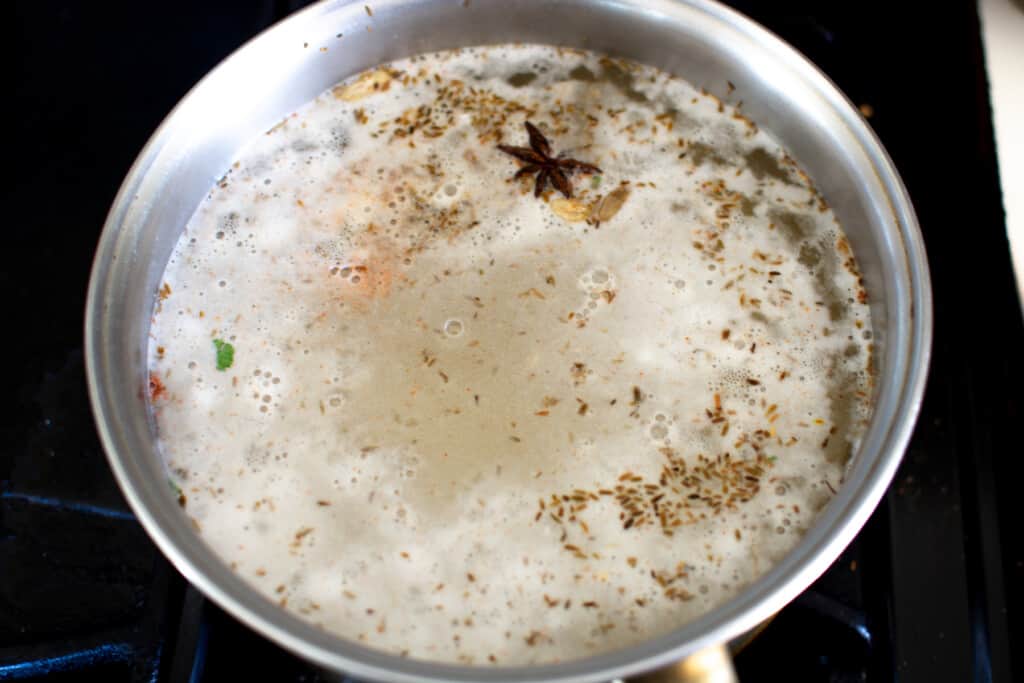 A metal saucepan on a stove top filled with water with spices floating on top. 