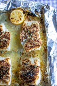 A baking sheet covered in foil with four pieces of broiled cod on it sitting on a purple checkered tablecloth.