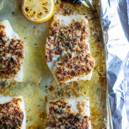 A baking sheet covered in foil with four pieces of broiled cod on it sitting on a purple checkered tablecloth.