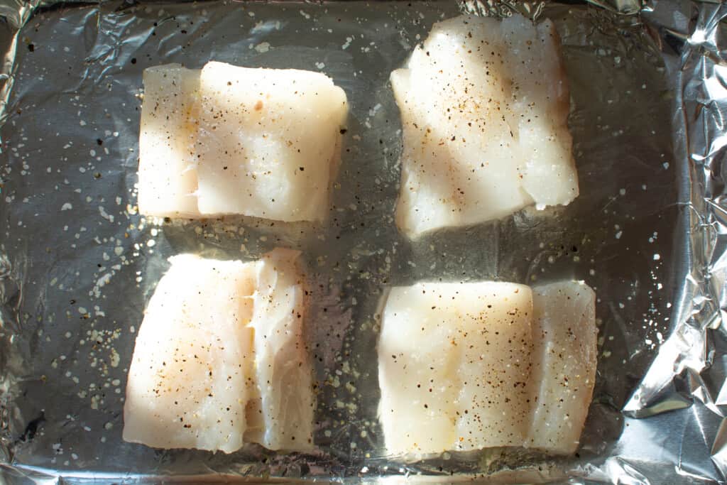 Four pieces of cod seasoned with salt and pepper sitting on a piece of aluminum foil on a baking sheet. 