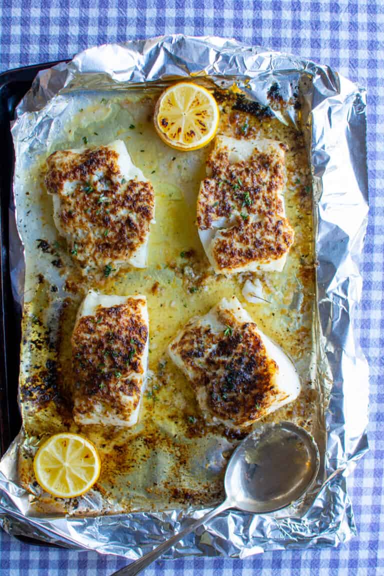 Four pieces of broiled cod sitting on a baking sheet lined with aluminum foil sitting on a purple checkered tablecloth.