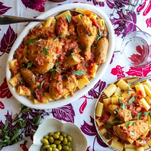 A table set with a big platter of chicken and pasta and another plate with sausage next to it.