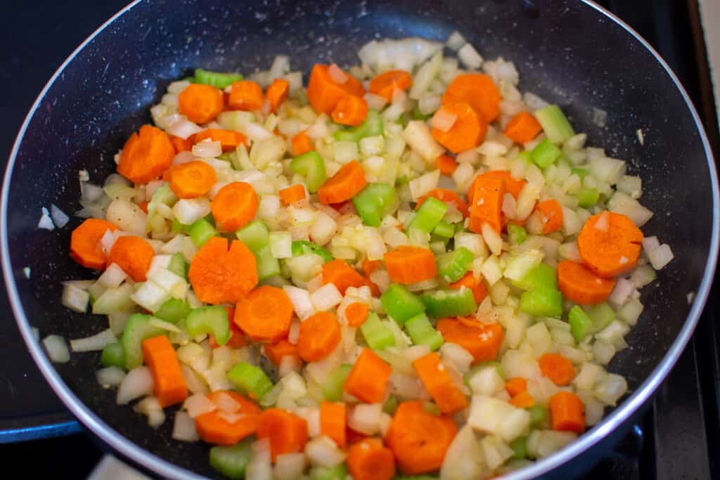 A black pan on the stove filled with onions, carrots, celery, and garlic sautéing. 