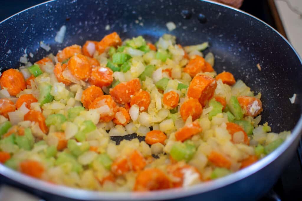 A black saute pan on the stove top with vegetables coated in flour sautéing on the stove. 