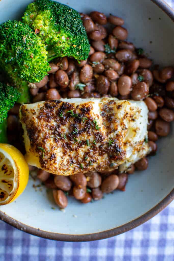 A piece of broiled cod sitting on top of brown beans next to steamed broccoli with a lemon wedge on one side. 