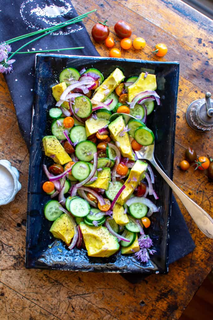 A black platter filled with cucumber tomato avocado salad on a wood table on a black napkin with cherry tomatoes around. 