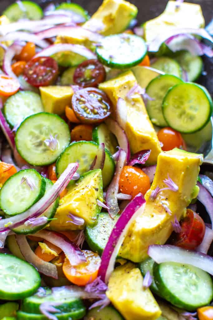 A close up of a cucumber tomato avocado salad with red onions and chive blossoms on top. 