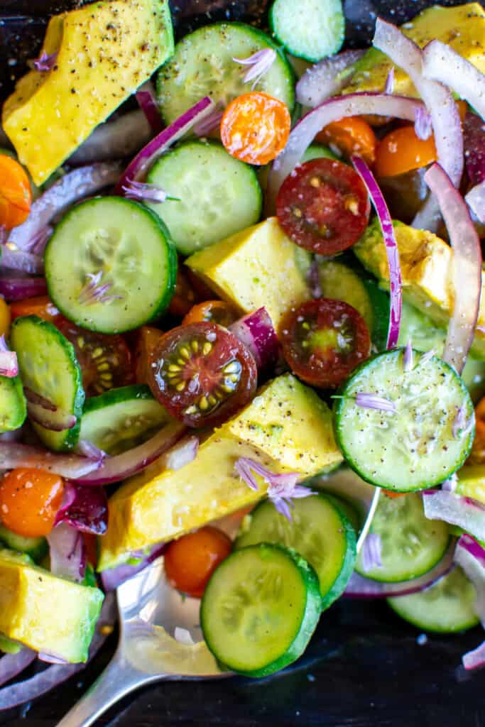 A cucumber salad with a serving spoon in the salad with cherry tomatoes, red onion, and avocado. 