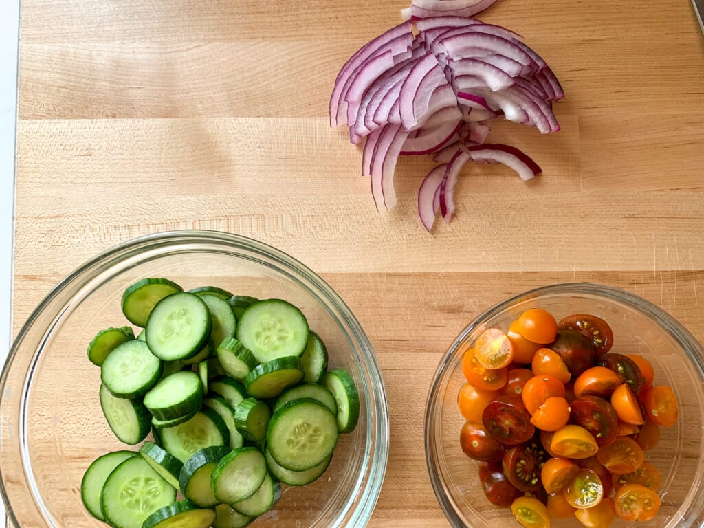 Cucumbers, red onion, and cherry tomatoes chopped on a wood counter. The cucumbers and tomatoes are in glass bowls. 