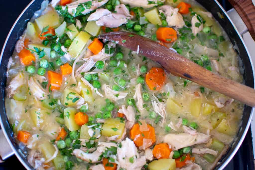 A black pan filled with shredded chicken, potatoes, frozen peas, parsley, carrots, celery, broth, and onions. 