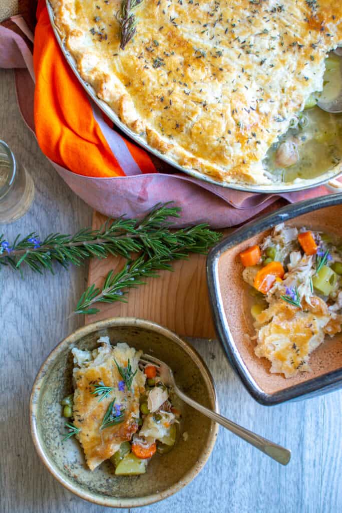 Two serving bowls with chicken pot pie in them next to a casserole dish of chicken pot pie and a couple sprigs of rosemary. 