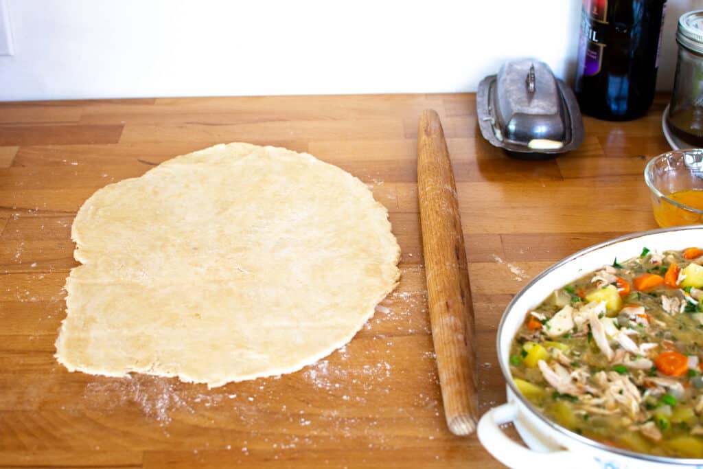 The pie crust for the dairy-free chicken pot pie being rolled out on a wood counter next to a casserole of filling. 