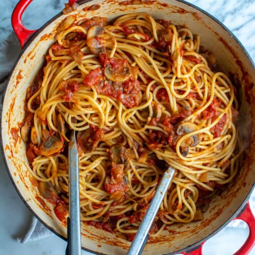 A red pot of mushroom spaghetti sauce with tongs sticking out of it.