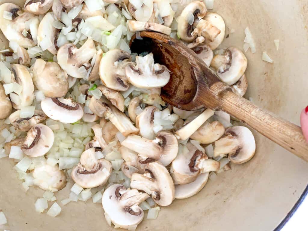 A wood spoon stirring mushrooms, onions, and garlic in a pot. 