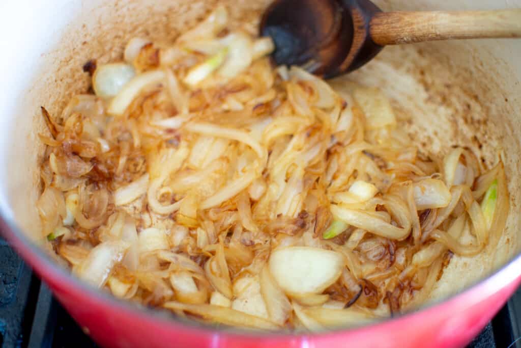 A red Dutch oven on a stove top with onions in it that are caramelizing with a wood spoon in the pot. 