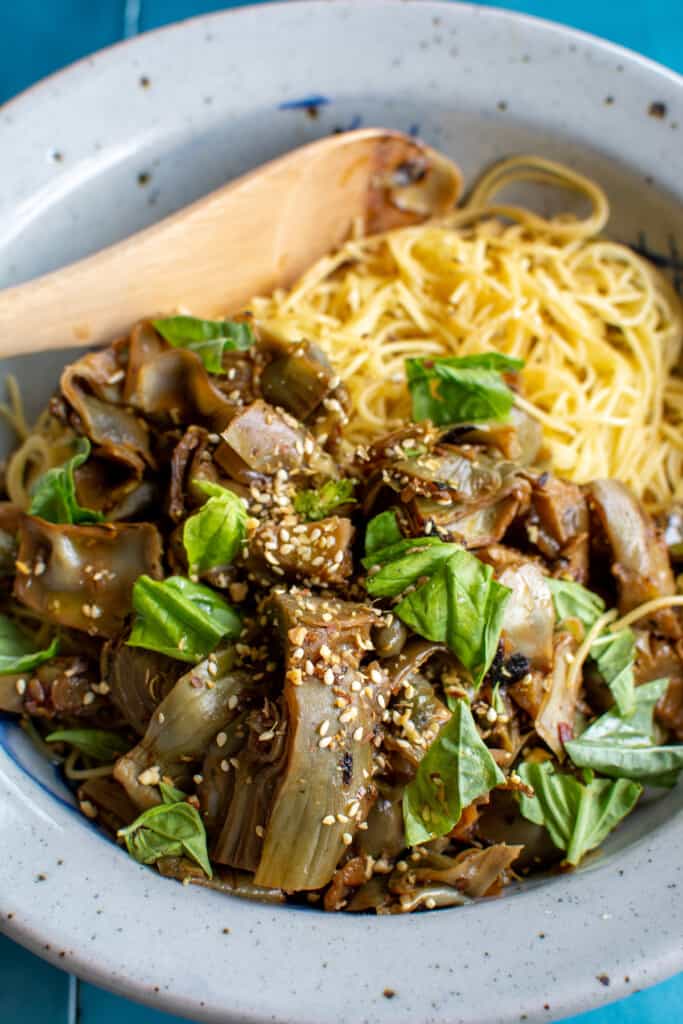 A bowl of angel hair pasta topped with braised artichokes and a wood spoon behind it with sesame seeds on top. 