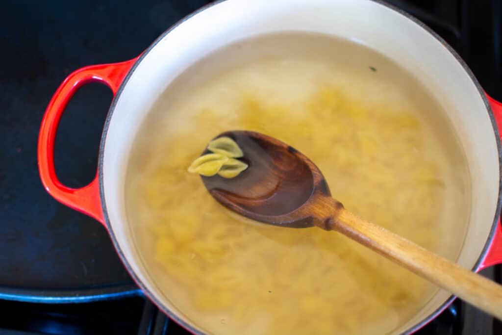 A red pot on a stove top filled with water and pasta cooking in the water with a wood spoon dipped in and holding pasta. 