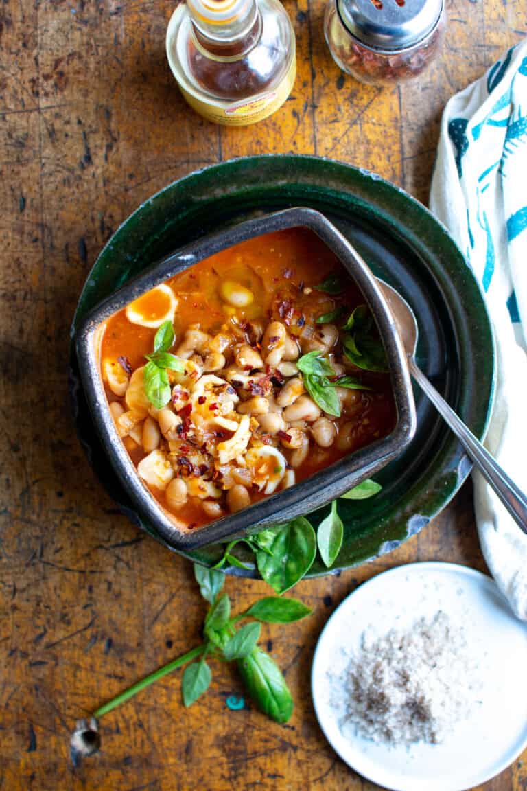 Overhead image of a bowl of pasta fagioli on a ceramic plate sitting on a wood table with fresh herbs on the side.
