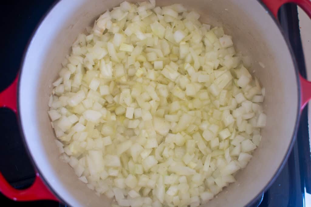 A red pot on the stove with chopped onions inside being sautéed. 