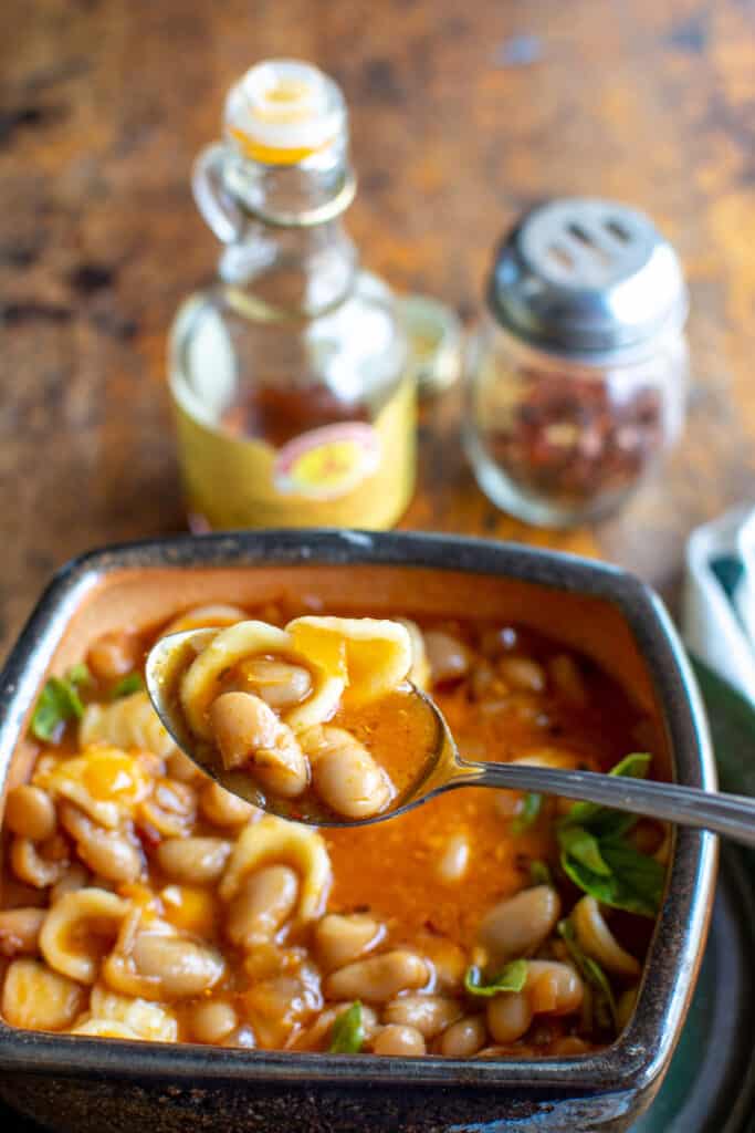 A bowl of pasta fagioli with a spoonful of the soup being held up to the camera with a jar of chili oil in the back. 