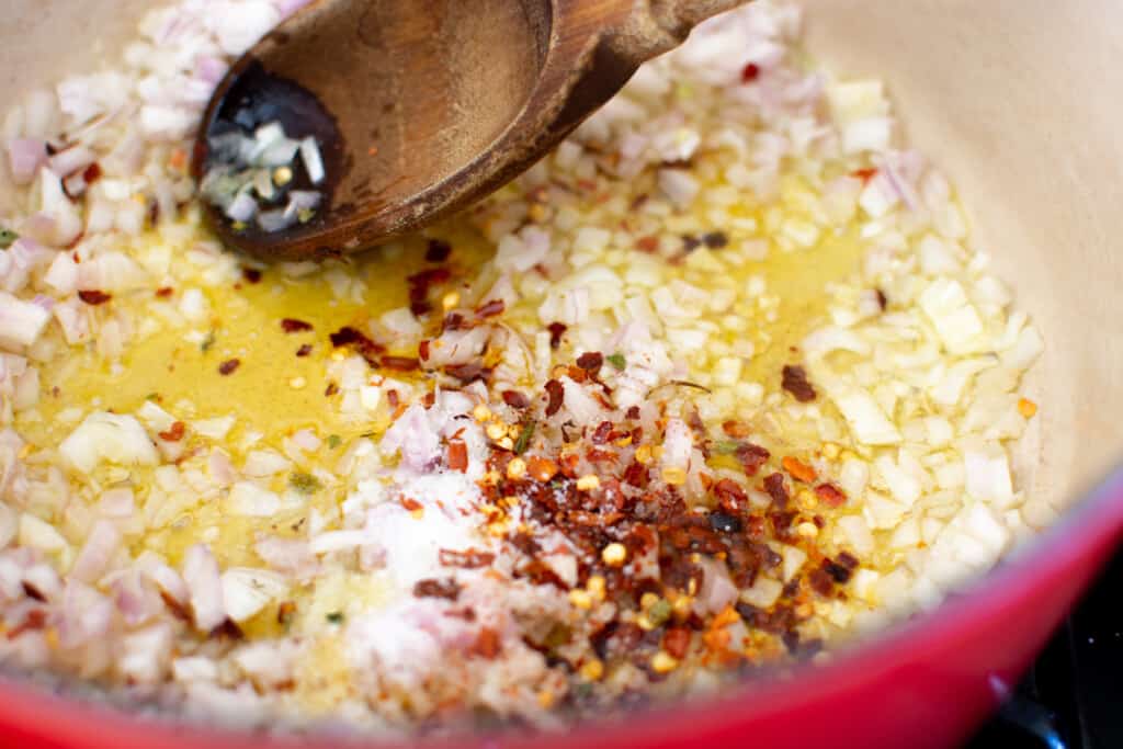 A pot with shallots and Chile flakes sautéing in olive oil with a wood spoon sticking out of one side of the pan. 