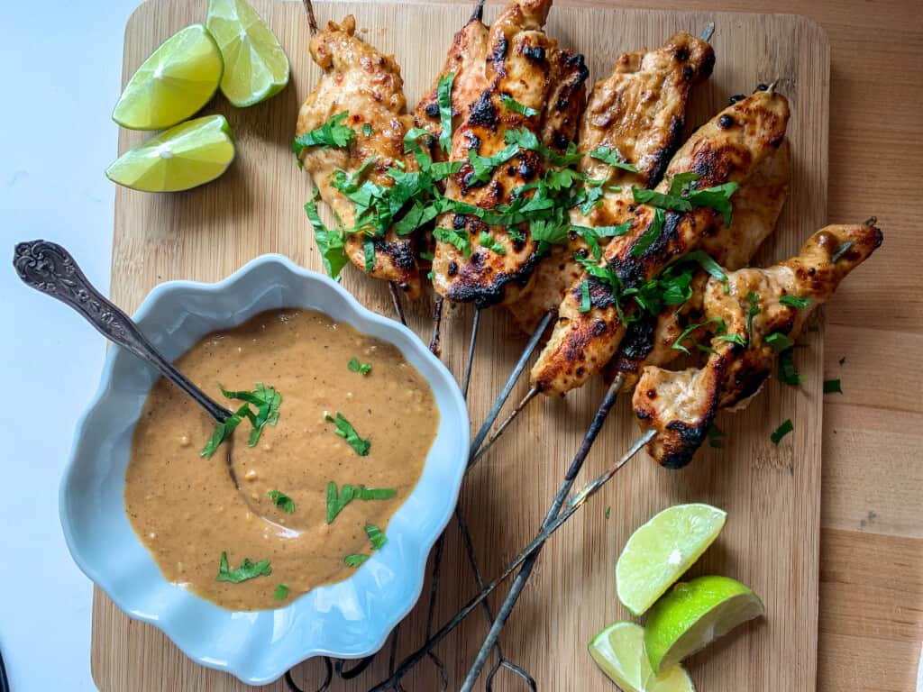 Several chicken skewers on a wood cutting board next to a bowl of sauce and limes. 