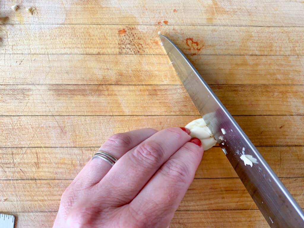 A hand chopping a clove of garlic on a wood cutting board. 