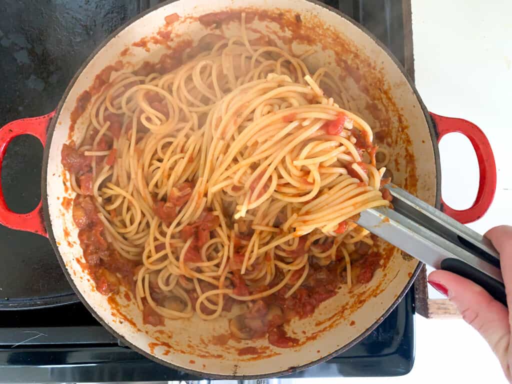 A red pot on the stove with a pair of tongs stirring spaghetti and mushroom sauce on the stove. 