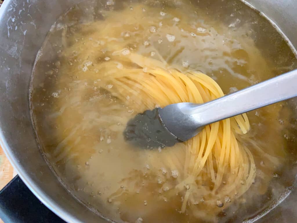 A metal pot with boiling water and spaghetti in the pot with a pair of tongs pulling it out of the water. 