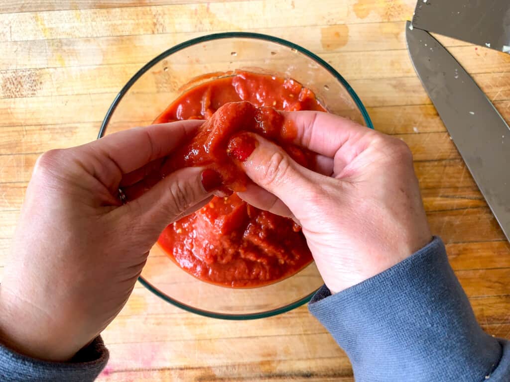 A hand crushing tomatoes over a glass container on a wood cutting board. 