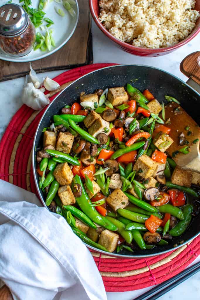 A black frying pan with tofu stir-fry in it sitting on a red mat next to a bowl of brown rice and a plate of scallions. 