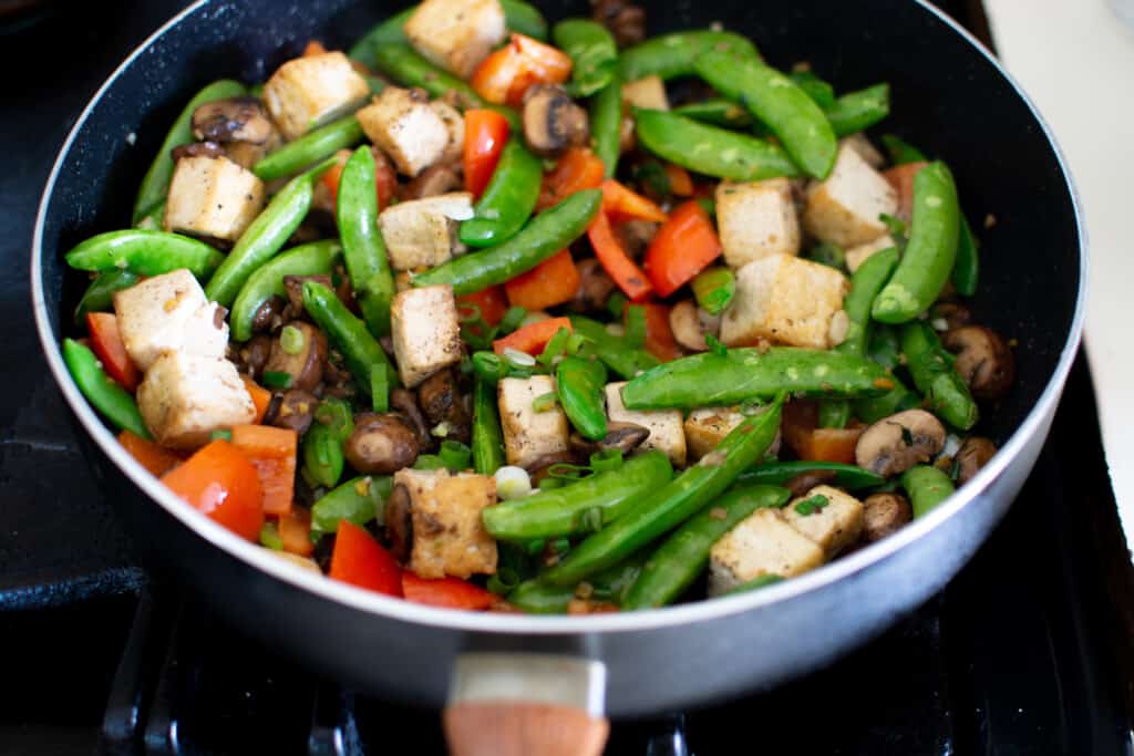 Tofu stir-fry in a pan with sugar snap peas, red bell peppers, pieces of tofu, mushrooms, and scallions. 