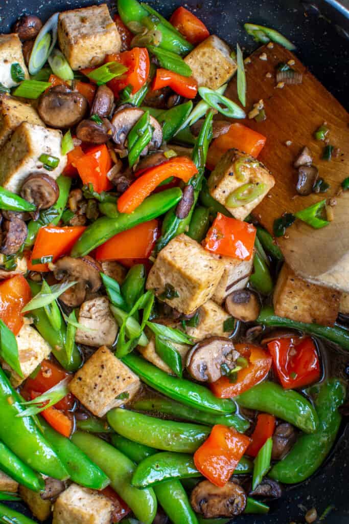 A close up image of tofu stir-fry with red bell peppers, sugar snap peas, thinly sliced scallions, and mushrooms. 