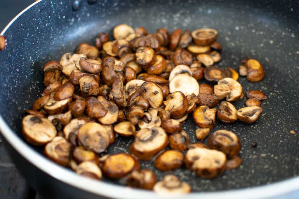 Crimini mushrooms in a black frying pan being sautéed in oil. 