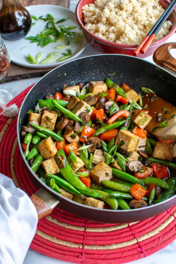 A black frying pan filled with tofu stir-fry on a red mat next to a plate of scallions and a bowl of rice.