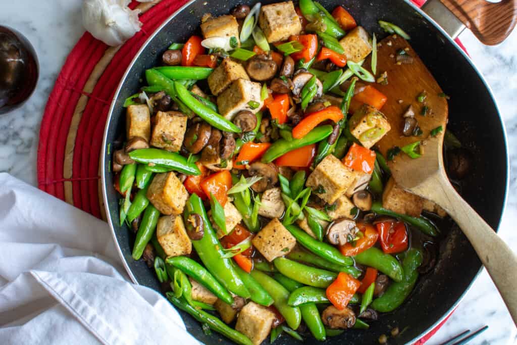 A black frying pan filled with tofu stir-fry sitting on a red mat with a wood spoon sticking out. 