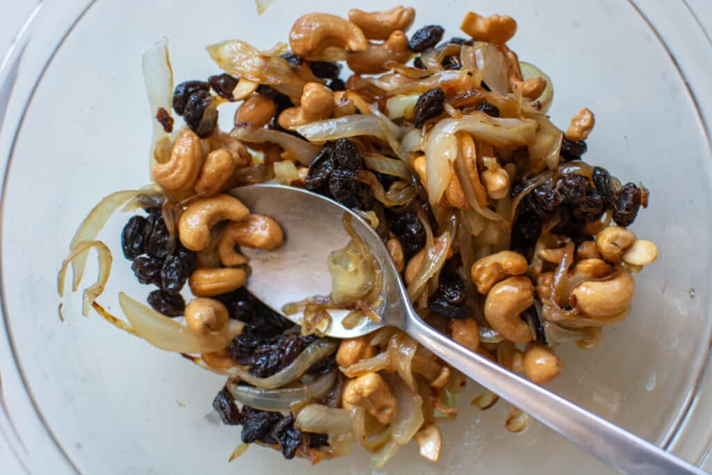 A glass bowl filled with cashews, raisins, caramelized onions and a spoon sitting in the bowl. 