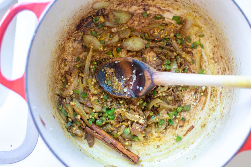 A red pot  with caramelized onions, jalapenos, cinnamon stick, and a wood spoon in the pot. 