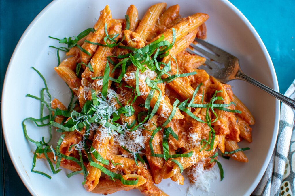 A white bowl of pasta with vodka sauce and thinly sliced basil and parmesan cheese and a fork sticking in the plate. 